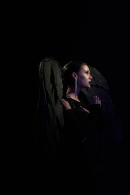 Kara kanatlı düşmüş melek kostümlü bir kadının yan görüntüsü. Karanlıkta dua ediyor, siyah fon.