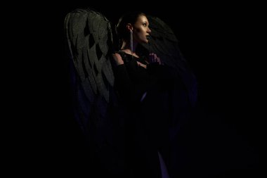 side view of mysterious woman in costume of demonic winged creature praying on black backdrop clipart