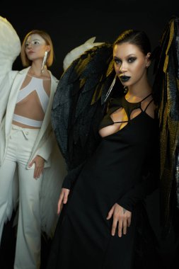 demonic woman looking at camera near angel on black backdrop, women in costumes of winged creatures clipart