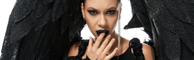 portrait of seductive woman in dark makeup and costume of black demon touching lips on white, banner clipart