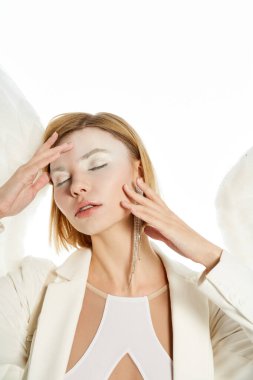 graceful woman in costume of winged angel posing with closed eyes and hands near face on white clipart