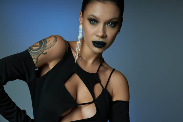 tattooed woman in shiny earring and sexy dress looking at camera on blue and grey, sinister beauty