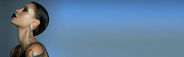 профиль татуированной женщины с готическим макияжем и блестящей серёжкой на синем сером фоне, баннер