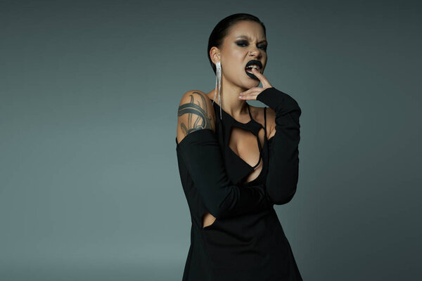 tattooed woman in black halloween dress, with wicked face expression looking at camera on grey