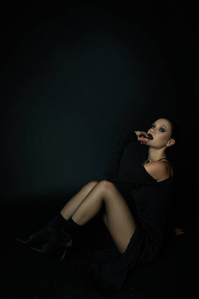 sensual woman in sexy halloween dress and dark makeup looking at camera while sitting on black