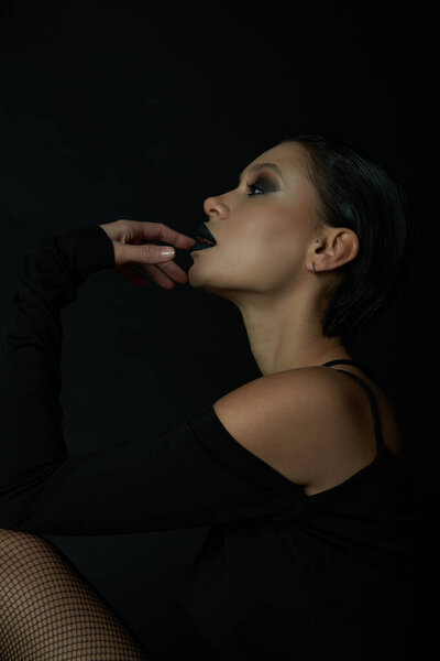 Gothic glamour, profile of enchanting woman with eerie makeup touching dark lips on black backdrop