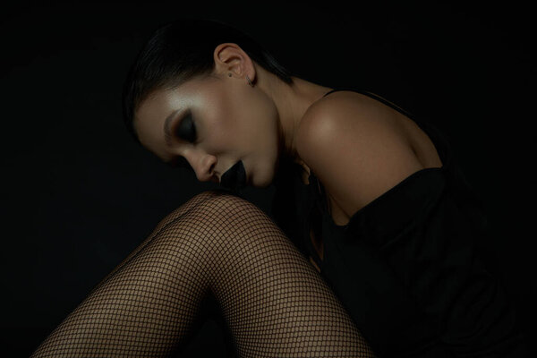 Gothic glamour, stylish woman with closed eyes and dark makeup sitting in fishnet tights on black