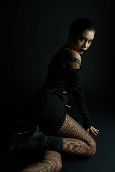passionate woman in sexy dress and fishnet tights sitting and looking at camera on black backdrop