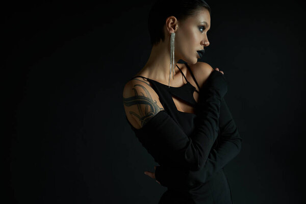 tattooed and glamour woman with eerie makeup posing in sexy dress on black, halloween concept