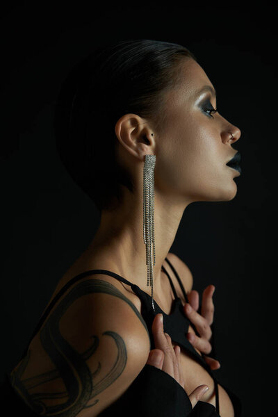 profile of sensual tattooed woman with dark makeup and shiny earring on black, halloween concept