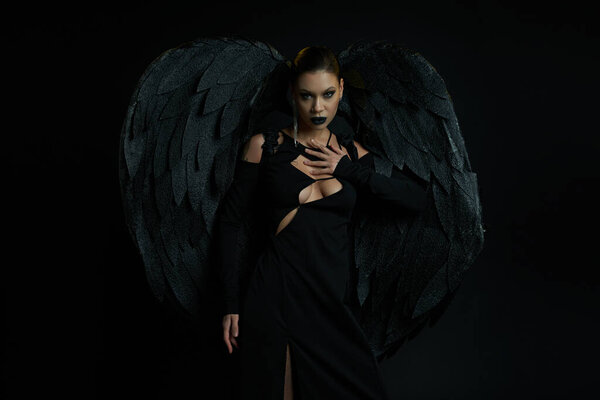 sexy woman in costume of fallen angel with dark wings looking at camera on black, halloween concept