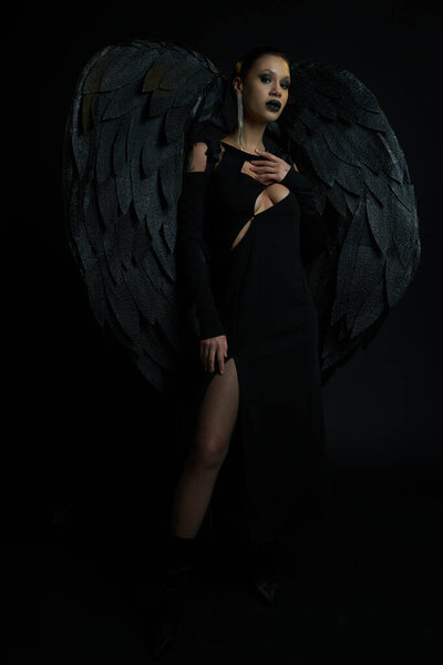 woman in sexy dress and fantasy dark demon wings looking at camera on black, halloween concept