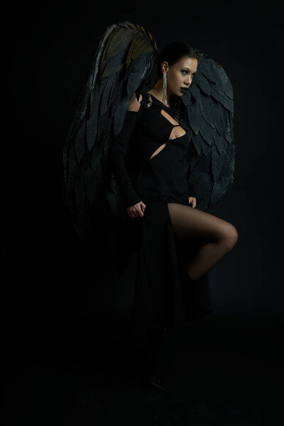 seductive woman in dark makeup and costume with demonic wings looking away on black, Halloween