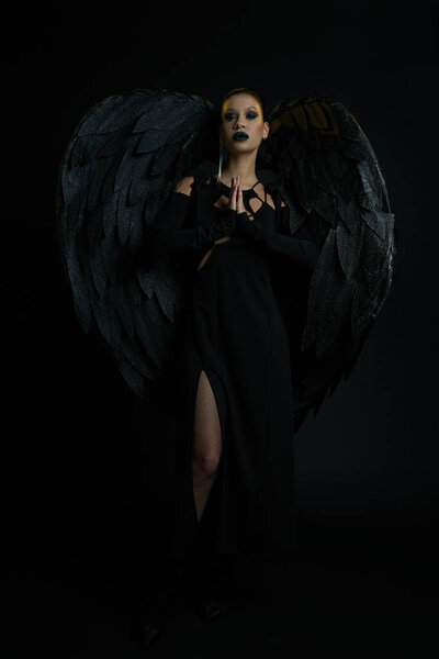 таинственная женщина в костюме крылатого существа, стоящего с молитвенными руками на черной, демонической красоте