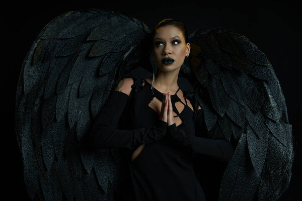 сексуальная женщина в костюме темного падшего ангела, стоящего с молитвенными руками и смотрящего на черное