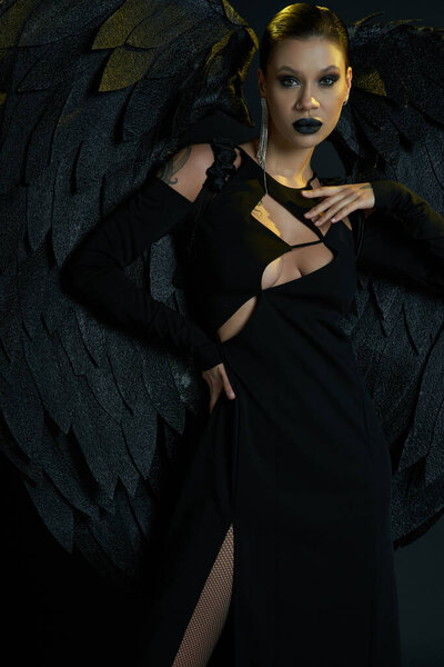 gothic beauty, tattooed woman in costume of dark winged creature standing with hand on hip on black