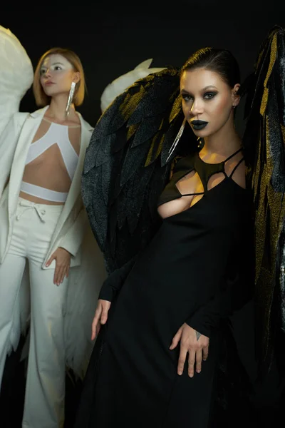 恶魔般的女人在黑暗的背景下看着镜头旁边的天使 穿着翅膀生物服装的女人 — 图库照片