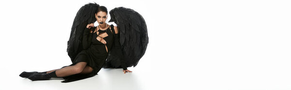 сексуальная женщина в темном макияже и черном костюме крылатого демона сидит на белом, баннер
