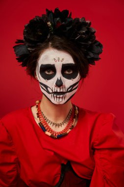 woman in dia de los muertos makeup and black wreath with colorful beads looking at camera on red clipart