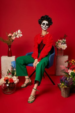 dia de los muertos makyajlı bir kadın geleneksel ofrenda yakınlarında kırmızı çiçekli bir koltukta oturuyor.