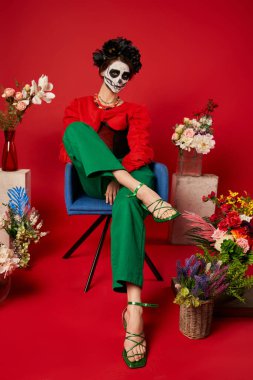 dia de los muertos makyajlı bir kadın geleneksel sunağın yanındaki koltukta oturuyor kırmızıda çiçeklerle