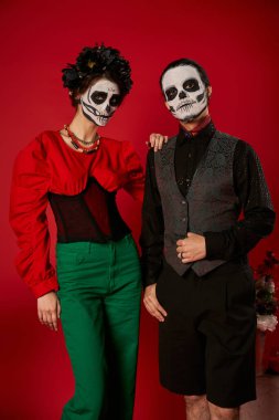 elegant couple in dia de los muertos makeup and festive attire looking at camera near flowers on red clipart