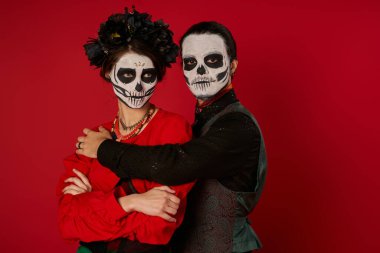 Lydia de los Muertos makyajlı bir çift, kırmızı kollarını kavuşturmuş kadını kucaklayan bir adam.