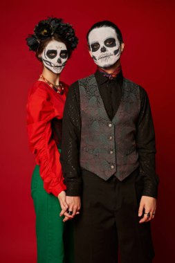 stylish couple in dia de los muertos sugar skull makeup holding hands and looking at camera on red clipart