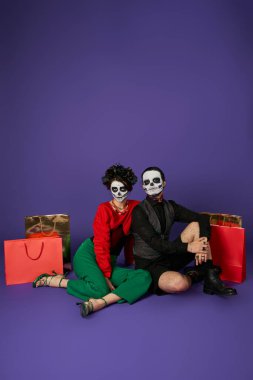 dia de los muertos couple in scary skeleton makeup sitting near shopping bags on blue, full length clipart