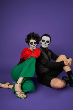 Los Muertos çifti. Kafatası makyajlı kadın. Mavi üzerine otururken ürkütücü adama yaslanıyor.