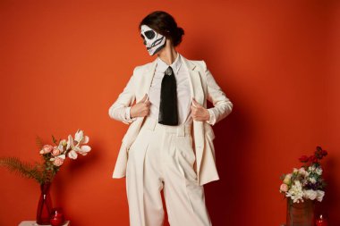 woman in catrina makeup and white suit near traditional dia de los muertos altar with flowers on red clipart