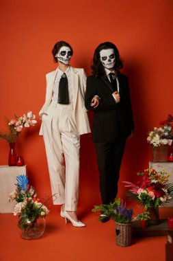 Catrina makyajlı ve takım elbiseli çift kırmızıda çiçeklerle birlikte dia de los muertos ofrenda yakınlarında poz veriyor.