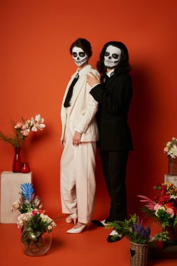 couple in skeleton makeup and suits near festive dia de los muertos ofrenda with flowers on red clipart