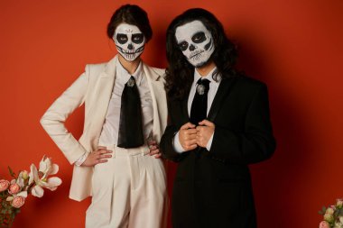 Catrina Calavera makyajlı zarif bir çift kırmızıda çiçek dekoru yakınındaki kameraya bakıyor.