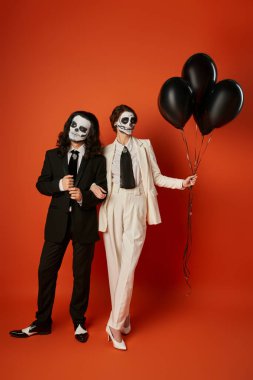 couple in skull makeup and festive attire near black balloons on red,   dia de los muertos party clipart