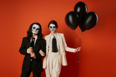 dia de los muertos party, elegant couple in sugar skull makeup standing with black balloons on red clipart