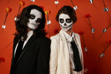 dia de los muertos 'un gözde çifti. Kafatası makyajı. Kırmızı arka planda çiçeklerle kameraya bakıyorlar.