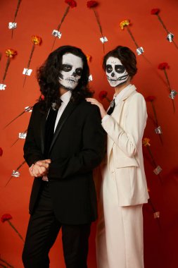 dia de los muertos catrina makyajlı kadın kırmızı arka planda karanfillerle erkeğin omzuna dokunuyor