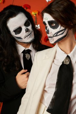 Los Muertos çifti, ürkütücü adam kırmızı arka planda çiçekli kafatası makyajı yapan kadına bakıyor.