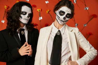 dia de los Muertos Catrina makyajlı bir kadın kırmızı çiçek arkaplanındaki korkunç adamın yanındaki kameraya bakıyor.