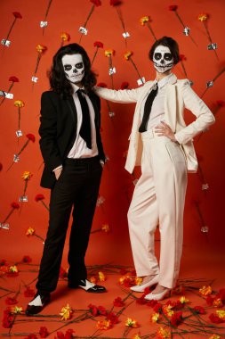 full length of stylish couple in skull makeup and suits standing on red backdrop with carnations clipart