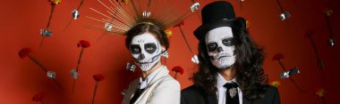 dia de los muertos makyaj ve şenlik kıyafetleri kırmızı arka planda çiçeklerle, afişlerle poz veren çift