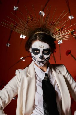 Didia de los Muertos 'taki kadın. Kafatası makyajı ve taç. Kırmızı çiçek arkaplanındaki kameraya bakıyor.