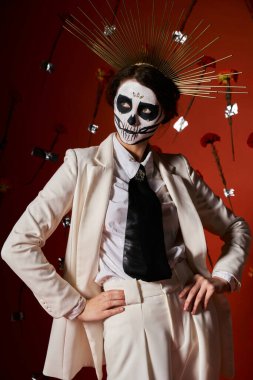 woman in dia de los muertos skull makeup and crown posing with hands on hips on red floral backdrop clipart