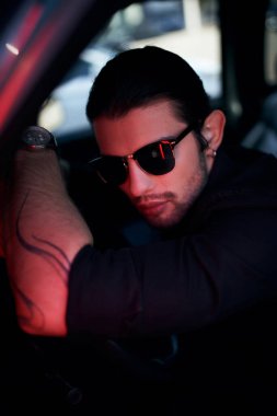 Araba direksiyonunda durup kameraya bakan dövmeli ve güneş gözlüklü seksi genç sürücü.