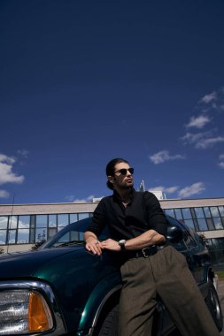 good looking young man posing next to his car wearing black shirt and brown pants and looking away clipart