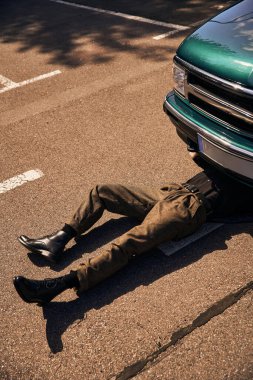 Siyah modaya uygun kentsel kıyafetli genç erkek model arabasının altında yatıyor ve onu tamir ediyor.