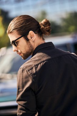 Güneş gözlüklü ve küpeli yakışıklı genç adam arabasının yanında poz veriyor, moda ve tarz.