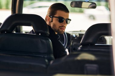 Direksiyon başında siyah kıyafetli yakışıklı bir sürücü ve kameraya bakıyor, moda konsepti.