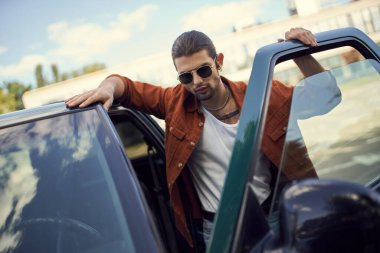 handsome sexy man with sunglasses and ponytail in vibrant outfit posing next to his car clipart